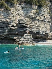 A lively group enjoys paddleboarding and swimming near stunning rocky cliffs, capturing the essence of a perfect beach day.