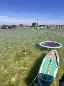 A child swims in clear waters near a paddleboard and floating mat, capturing the essence of fun water activities on a sunny day.