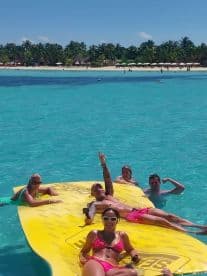 A lively group enjoys a sunny day on a floating mat in crystal-clear waters, perfect for beach fun and relaxation.