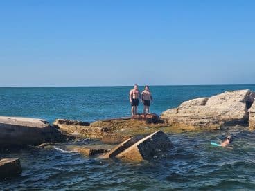 Two friends enjoy a sunny day at the beach, standing on rocks while another snorkels nearby — a perfect scene for summer adventures and water activities.