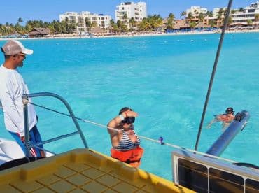 A child in a life jacket prepares to snorkel while an adult watches, with another swimmer in the background — a perfect family adventure in the clear blue waters.