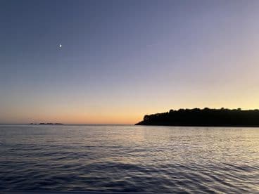 A serene sunset view over calm waters, showcasing the coastline and a crescent moon — perfect for tranquil evening boat rides.