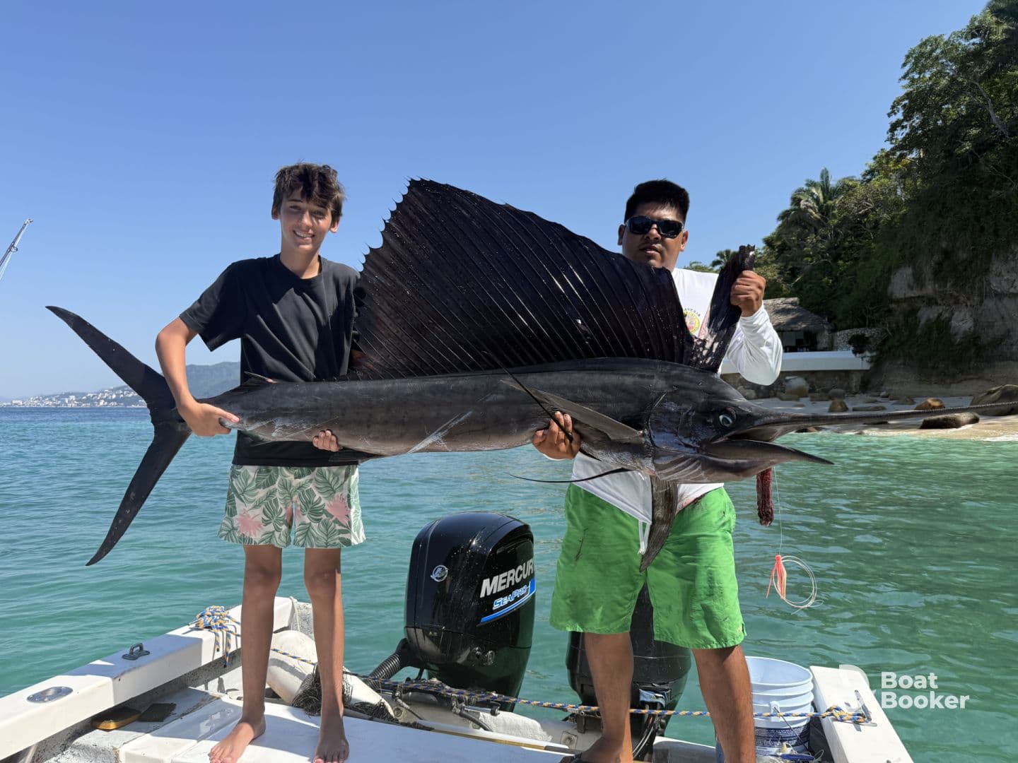 A young boy proudly holds a large sailfish alongside an adult on a fishing boat, showcasing a successful catch in beautiful coastal waters.