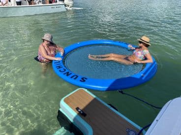 Two women enjoy a sunny day in the water, one relaxing on a floating mat while the other wades nearby — a perfect scene for summer fun and relaxation.