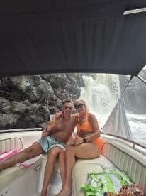 A couple enjoys refreshing drinks while relaxing on their boat near a stunning waterfall, capturing the essence of adventure and romance on the water.