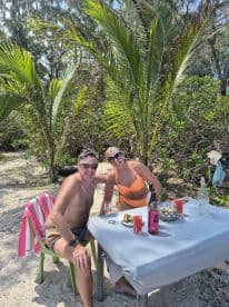 A cheerful couple enjoys drinks at a beachside table, surrounded by lush greenery — perfect for a relaxing getaway or romantic celebration.