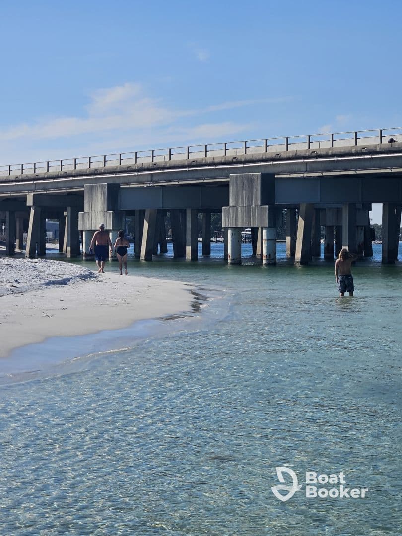 A couple strolls along the beach under a bridge, enjoying the clear waters and sandy shores — a perfect scene for a relaxing day by the water.