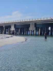 A couple strolls along the beach under a bridge, enjoying the clear waters and sandy shores — a perfect scene for a relaxing day by the water.