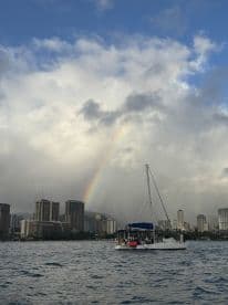A picturesque scene of a catamaran sailing near a vibrant rainbow and the skyline of Honolulu, capturing the beauty of a sailing adventure in Hawaii.
