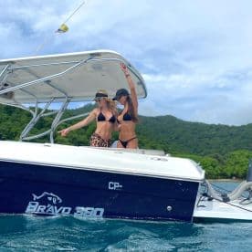 Two women celebrate on the deck of a Bravo 360 boat, enjoying a fun day on the water surrounded by lush green hills.