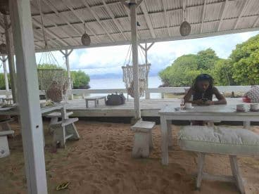 A person is seated at a sandy location under a covered area, seemingly engaged with their phone. The setting appears relaxed, but the overall context is unclear.
