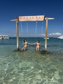 Two friends enjoy a playful moment on swings in shallow water at Mansa Bay, creating a perfect backdrop for fun beach memories.