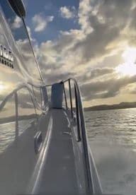 A sleek view of a boat's side as it glides through the water, showcasing the vessel's design against a backdrop of dramatic clouds and a shimmering sea.