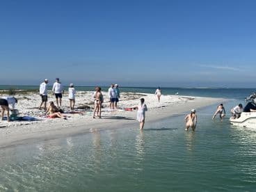 A lively group enjoys a sunny day at the beach, with some swimming and relaxing on towels, while a boat is anchored nearby — perfect for a fun day out on the water.