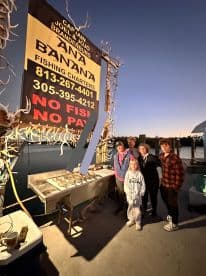 A family poses in front of the Banana Fishing Charters sign after a successful fishing trip, showcasing their catch and enjoying a memorable moment together.