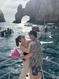 A couple shares a romantic moment aboard a boat in Cabo San Lucas, celebrating with a bouquet against the stunning backdrop of the iconic rock formations.