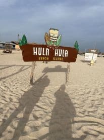 A sign for Hula Hula Beach Island is displayed on a sandy beach, but the image lacks clarity and detail.