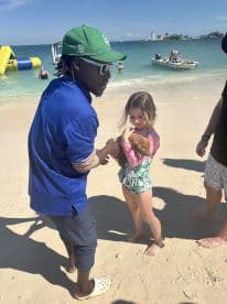 A young girl holds a piglet on the beach while interacting with a guide, showcasing a unique wildlife experience in a tropical setting.