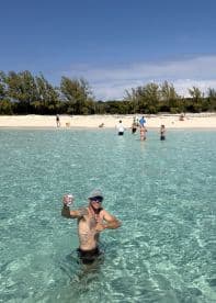 A man enjoys a refreshing drink while wading in crystal-clear waters at a beautiful beach, surrounded by friends and family — perfect for a fun day in the sun.