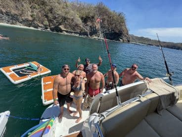 A lively group enjoys a fun day on the water, celebrating with drinks and fishing on a pontoon boat, surrounded by stunning coastal scenery.