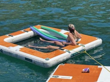 A woman relaxes on a floating mat in clear waters, enjoying the sun with pool noodles nearby — perfect for a day of leisure on the water.