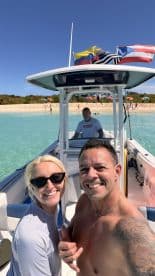 A cheerful couple takes a selfie on their boat with a beautiful beach backdrop, capturing the essence of a fun day on the water.