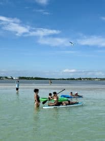A lively group enjoys paddleboarding and swimming in shallow waters, capturing the essence of a fun day at the beach.