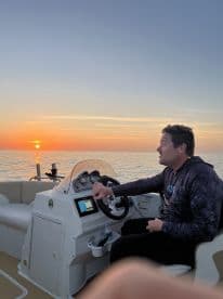 A man navigates a boat at sunset, creating a serene atmosphere perfect for evening cruises. The stunning sunset reflects on the water, enhancing the boating experience.