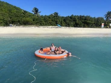 A joyful group enjoys a sunny day in an inflatable float, swimming in the clear waters near a beautiful beach — perfect for family fun and relaxation.