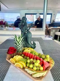 A beautifully arranged fruit platter featuring a variety of fresh fruits, complemented by a decorative mermaid statue, perfect for onboard catering during luxury yacht charters.