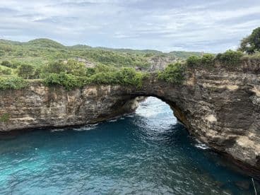 A stunning view of a natural rock arch along the coastline, surrounded by lush greenery and clear blue waters — a perfect destination for sightseeing and exploration.