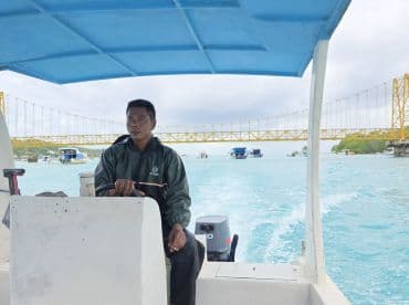 A boat driver navigates through turquoise waters with a striking yellow bridge in the background, showcasing the beauty of the surroundings.