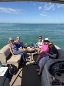 A cheerful group enjoys a relaxing day on a pontoon boat, sharing food and drinks while taking in the beautiful water views — perfect for a fun outing with friends.