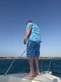 A man stands on the bow of a boat, fishing in the open ocean, showcasing a vibrant summer outfit and a beautiful coastal backdrop.