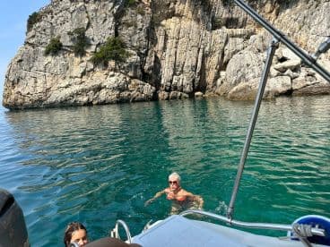 A woman swims in crystal-clear waters near a rocky coastline, while a child enjoys the refreshing sea — a perfect snapshot of a fun day on the water.