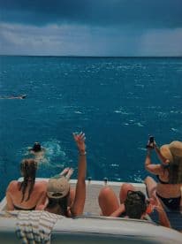 A lively group enjoys a fun day on the water, with one person swimming and others relaxing on the boat, capturing the moment with their phones.