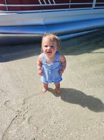 A cheerful child stands on the sandy shore near a pontoon boat, capturing a joyful moment during a family outing on the water.