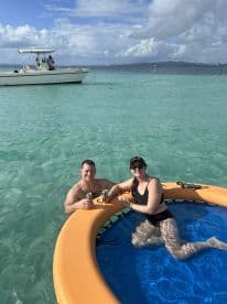 A couple enjoys refreshing drinks while relaxing on a floating mat in crystal-clear waters, perfect for a fun day on the water.