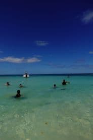 A lively scene of families enjoying a day at the beach, swimming in clear turquoise waters with a boat anchored nearby — perfect for summer fun and water activities.