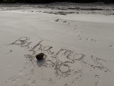 A sandy beach scene featuring the words 'Bahamas 2026' written in the sand, accompanied by a coconut. A serene setting ideal for beach lovers.