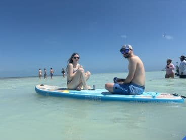 A couple enjoys paddleboarding in shallow waters, sipping drinks while surrounded by friends at the beach — a perfect day for fun and relaxation.
