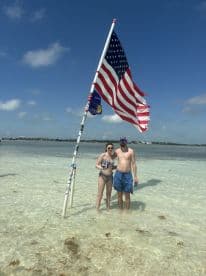 A couple enjoys a sunny day at the beach, standing in shallow water beside an American flag — a perfect moment for summer celebrations and beach outings.