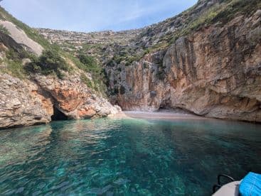 A stunning view of a secluded beach with crystal-clear waters and a cave, featuring swimmers enjoying the serene surroundings — an ideal spot for exploration and relaxation.