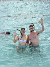 A couple enjoys snorkeling in crystal-clear waters, waving for the camera while a third person swims nearby — a perfect snapshot of underwater adventure.