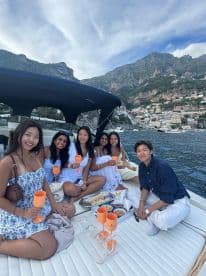 A joyful group of friends enjoys a celebration on a yacht, complete with cocktails and snacks, set against a stunning coastal backdrop.