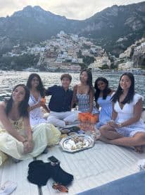 A joyful group of friends enjoys a scenic boat outing with delicious food and drinks, set against the stunning backdrop of Positano's picturesque coastline.