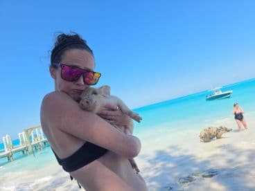 A woman holds a piglet while enjoying a beach day, with another pig wandering in the background — a delightful encounter with the famous swimming pigs.