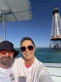 A couple takes a cheerful selfie on a boat with a lighthouse in the background, capturing a memorable moment on the water.