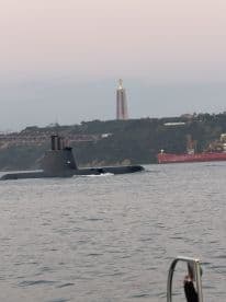 A submarine navigates through the water with a scenic backdrop featuring a prominent statue and bridge, showcasing a unique maritime experience.
