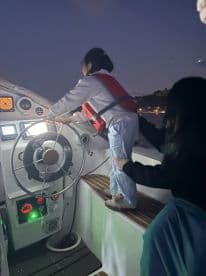 A young girl, wearing a life jacket, stands at the helm of a boat, guided by an adult, showcasing a fun and educational boating experience for children.